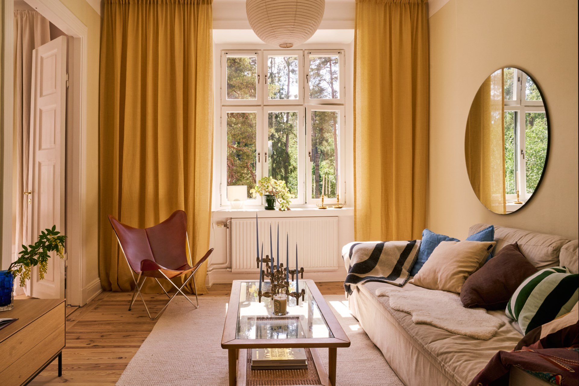 Helles Wohnzimmer mit goldgelben Vorhängen, beigem Sofa mit bunten Kissen, rotem Ledersessel und Couchtisch aus Glas.