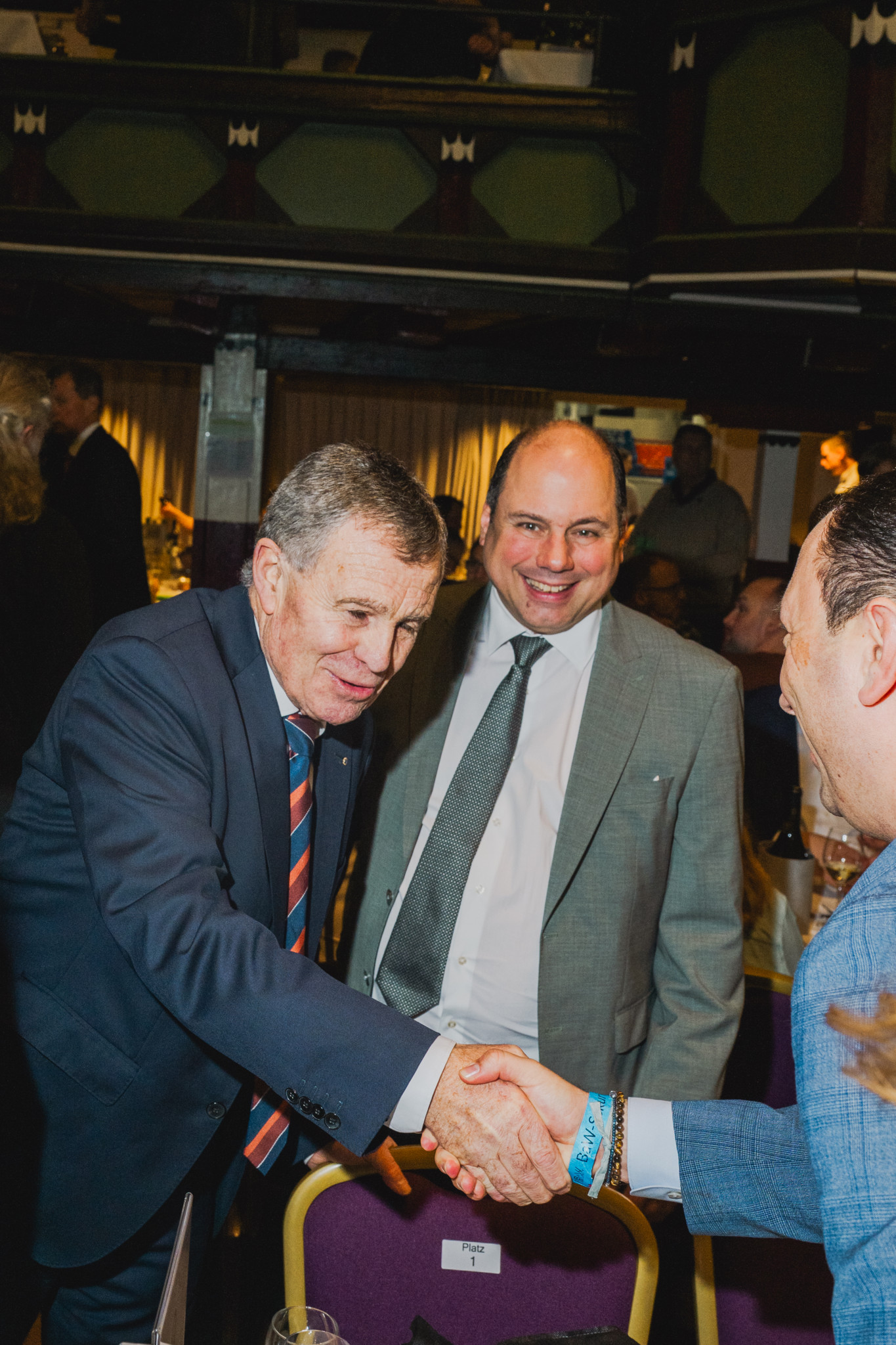 Drei Männer in Anzügen schütteln sich die Hände in einem formellen Veranstaltungsraum.