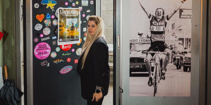 Frau mit langen blonden Haaren steht in einer Türöffnung, umgeben von Postern und Aufklebern, darunter ein Bild von einem Radrennfahrer.