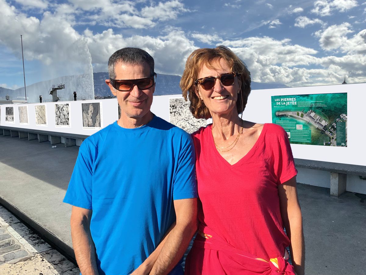 Deux personnes souriantes, un homme en t-shirt bleu et une femme en t-shirt rouge, posent devant une exposition de photos en plein air et un jet d'eau.