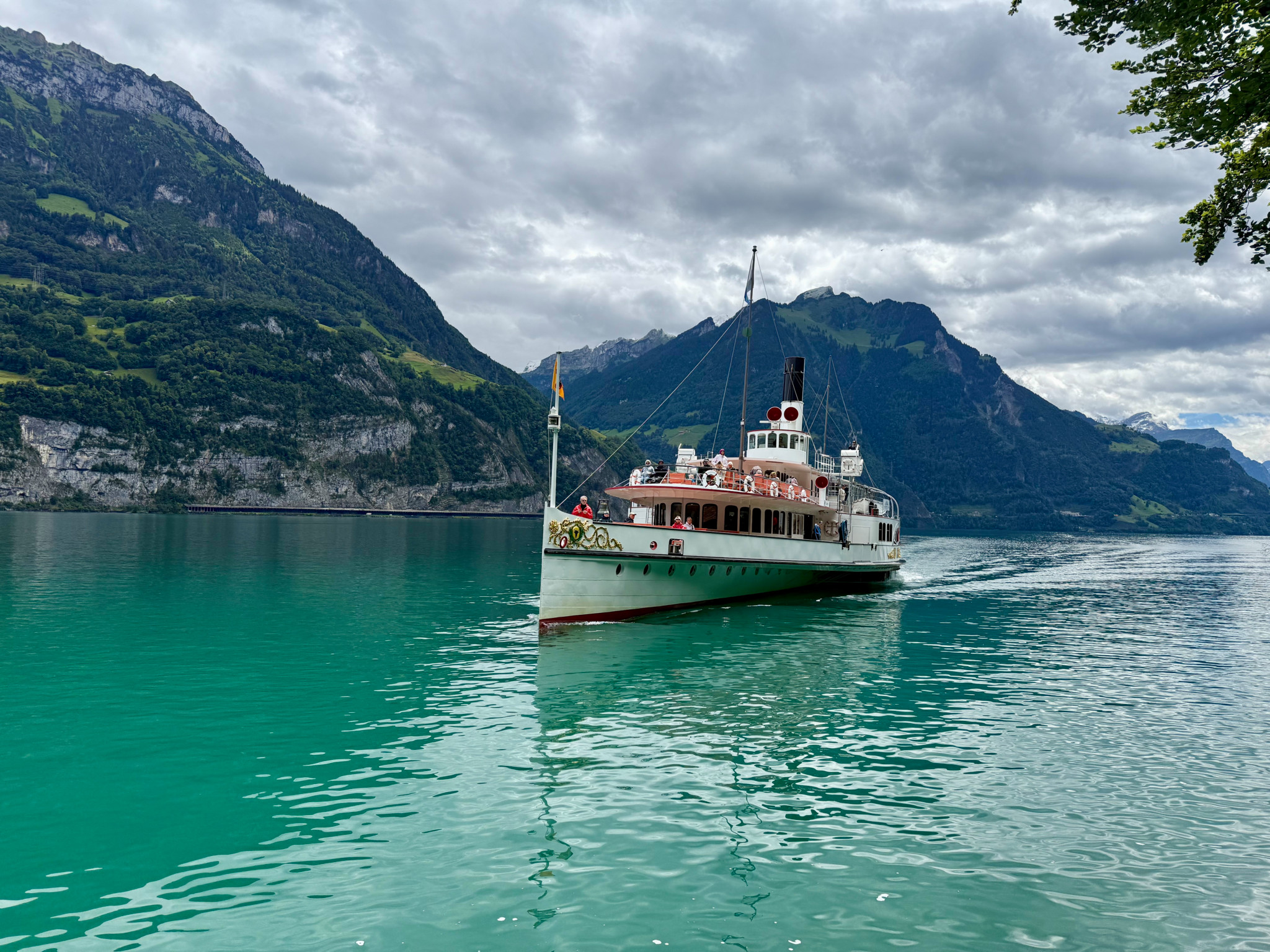 Historisches Dampfschiff auf einem türkisfarbenen Schweizer See, umgeben von bewaldeten Bergen unter bewölktem Himmel.