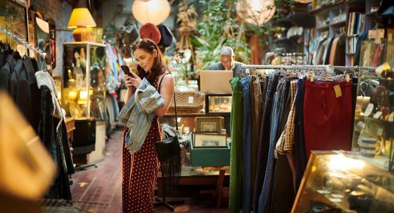 Une femme examine un vêtement dans une boutique vintage remplie de vêtements suspendus et de divers objets anciens.