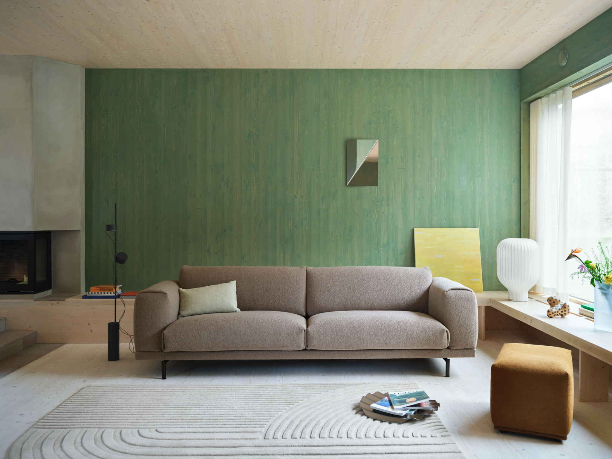 Modernes Wohnzimmer mit beigem Sofa vor grüner Wand, Kamin, weissem Teppich und gelbem Hocker am Fenster
