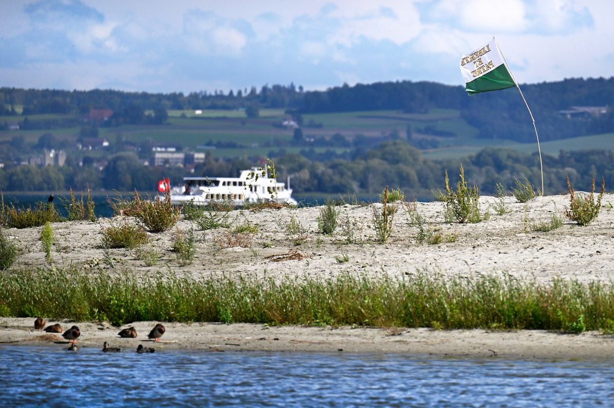 Un îlot avec un drapeau vaudois planté au centre apparaît après les travaux de revitalisation à l'embouchure de la Broye dans le lac de Morat, avec un bateau en arrière-plan.