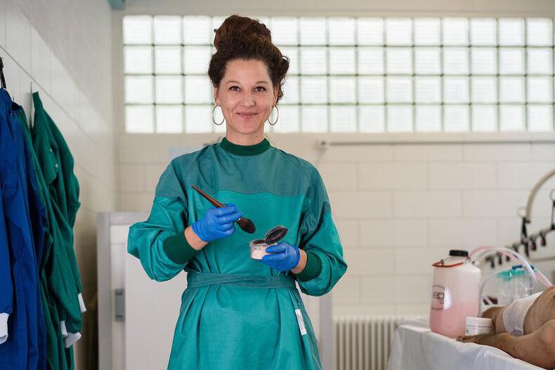 Myriame Marti souriante en blouse verte et gants bleus tient des ustensiles de travail dans une pièce blanche.