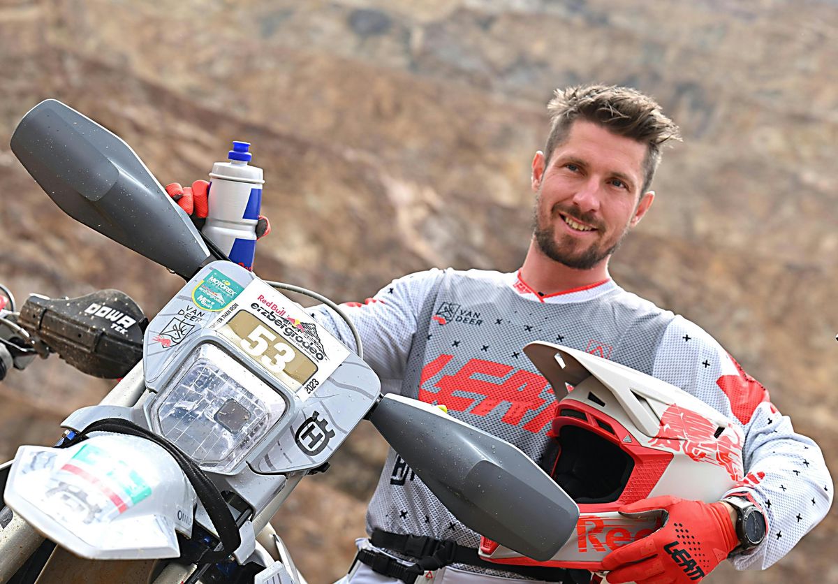 Motocycliste souriant en tenue de course, tenant un casque rouge à côté de sa moto Husqvarna, avant le Red Bull Hare Scramble 2023 en Autriche.