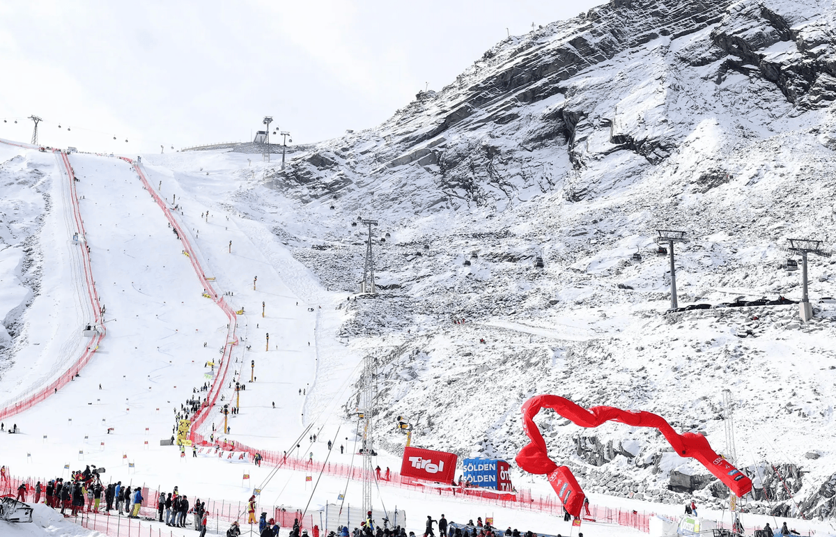 Piste de ski enneigée avec des skieurs et des spectateurs au pied de la montagne, entourée de télésièges et de bannières rouges.