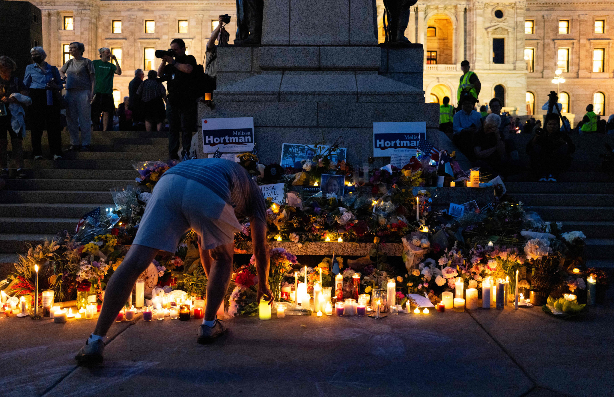 Eine Person legt eine Kerze an einem Gedenkort vor dem Minnesota State Capitol in St. Paul nieder, umgeben von Blumen und Kerzen in der Abenddämmerung.