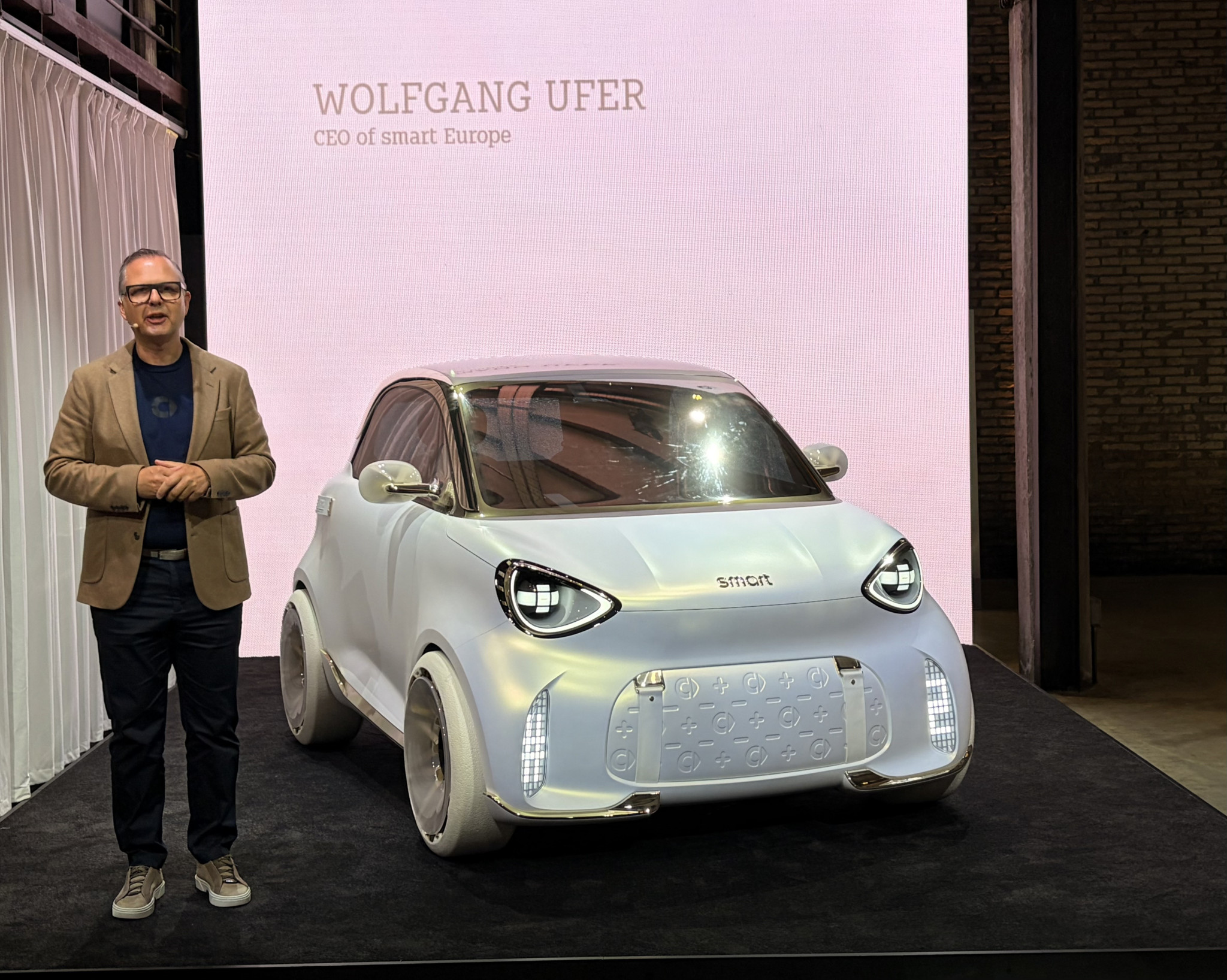Wolfgang Ufer, CEO von Smart Europe, steht neben einem Smart Elektroauto-Konzeptfahrzeug auf einer Präsentationsbühne.
