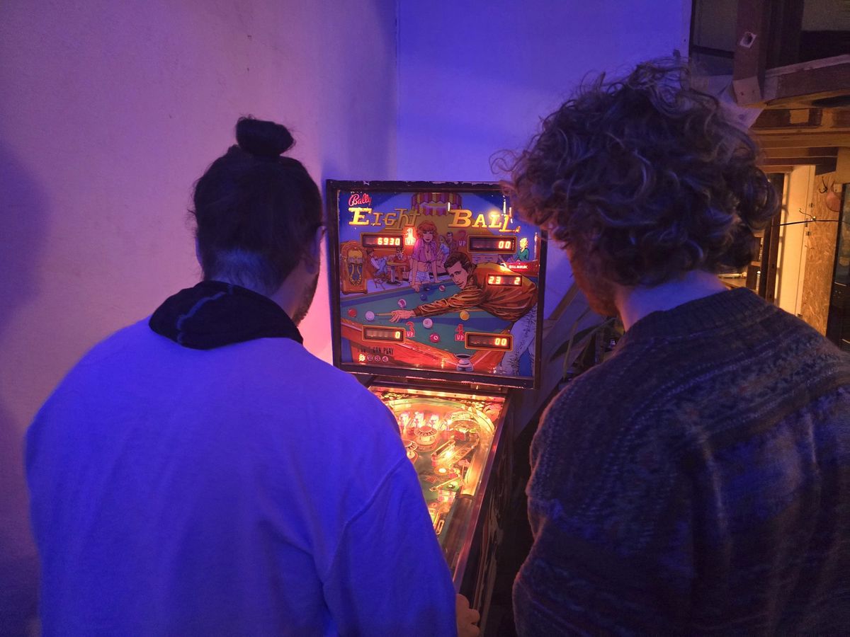 Deux personnes jouent à un flipper rétro, éclairé dans une ambiance chaleureuse et tamisée.