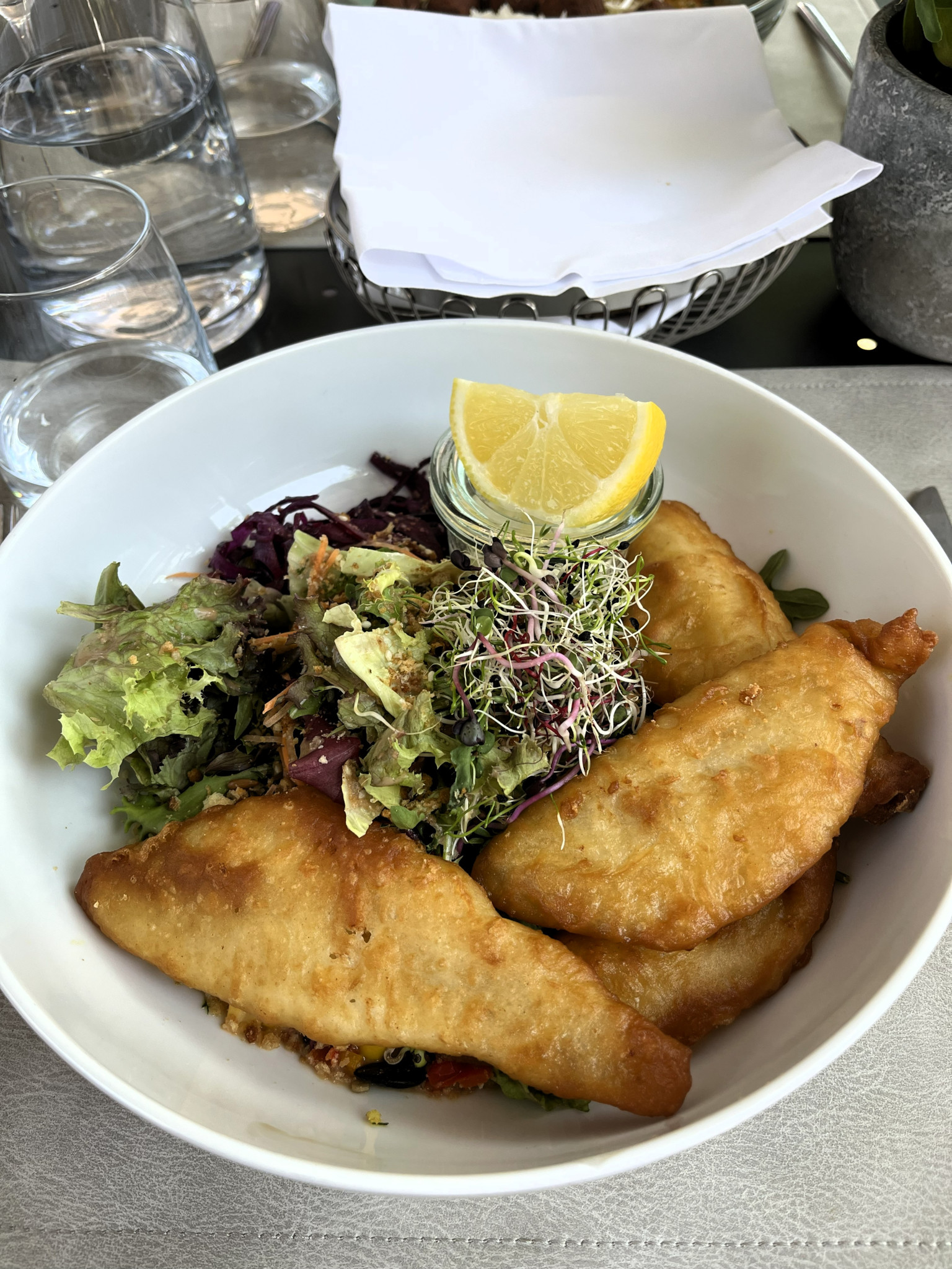Gebratenes Fischfilet im Teigmantel mit gemischtem Salat, Sprossen und Zitronenschnitz in einem weissen tiefen Teller.
