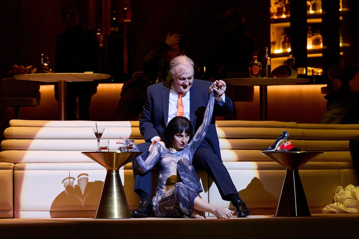 Scène d’un spectacle avec un homme en costume tenant la main d’une femme allongée sur un canapé, entourés de tables avec des verres.