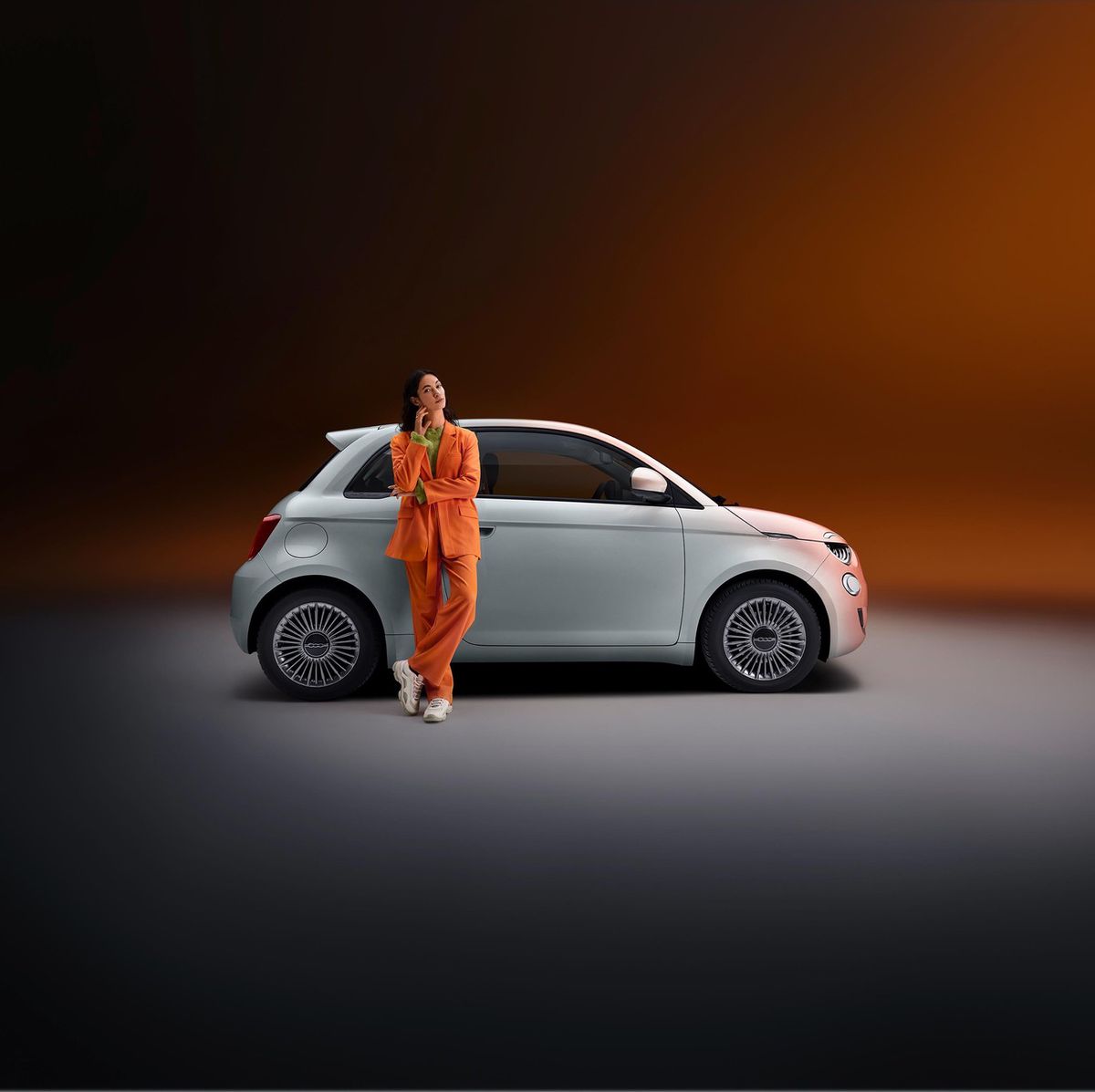 Personne en costume orange posée contre une petite voiture blanche sur un fond dégradé orange sombre.