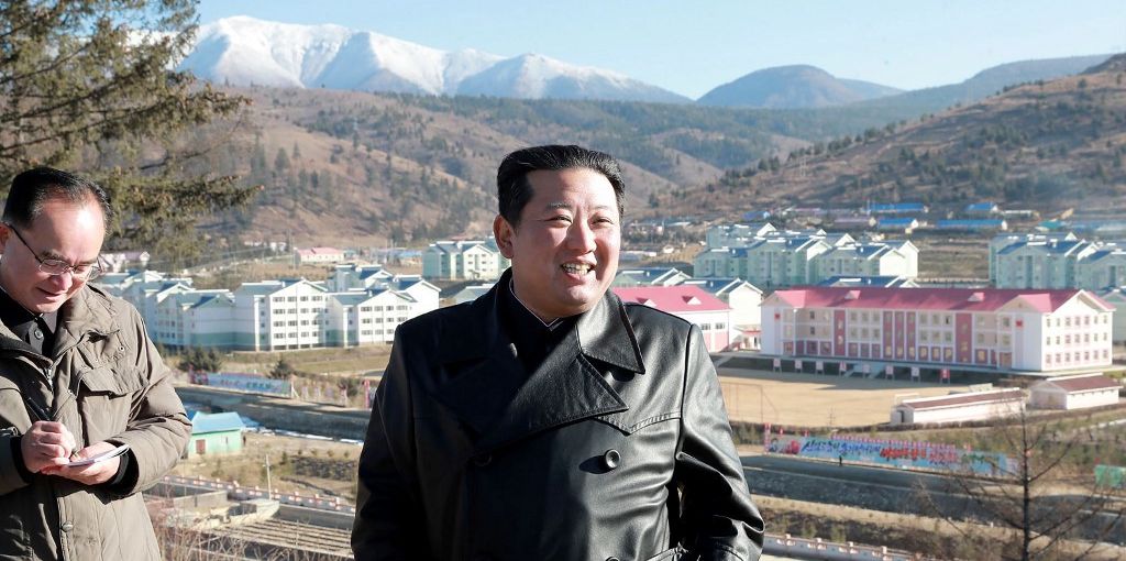 Ein Mann in einem schwarzen Leder-Mantel lächelt, während er in eine bergige Umgebung mit Gebäuden im Hintergrund schaut.