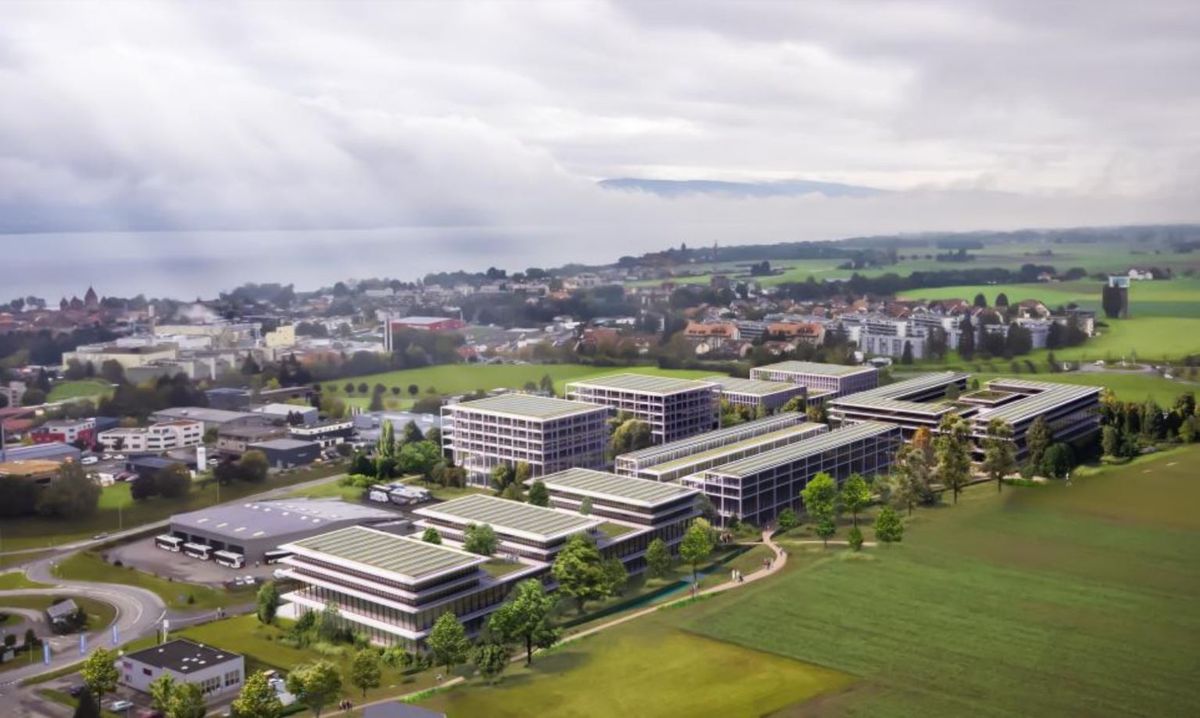 Vue aérienne d'un complexe de bâtiments modernes entouré de verdure et de champs, avec un arrière-plan de ville et de lac.