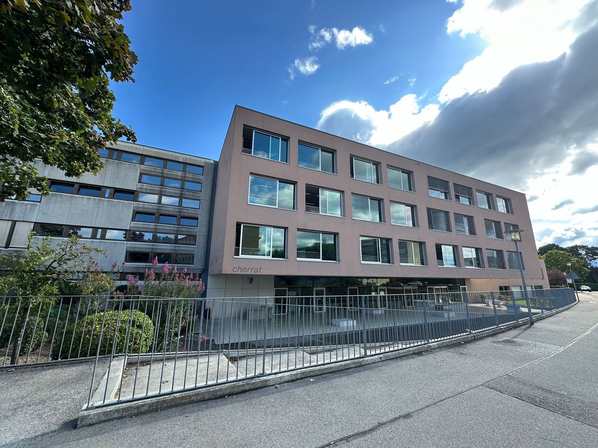 Bâtiment moderne à trois étages avec de grandes fenêtres et un arbre à gauche, sous un ciel partiellement nuageux.