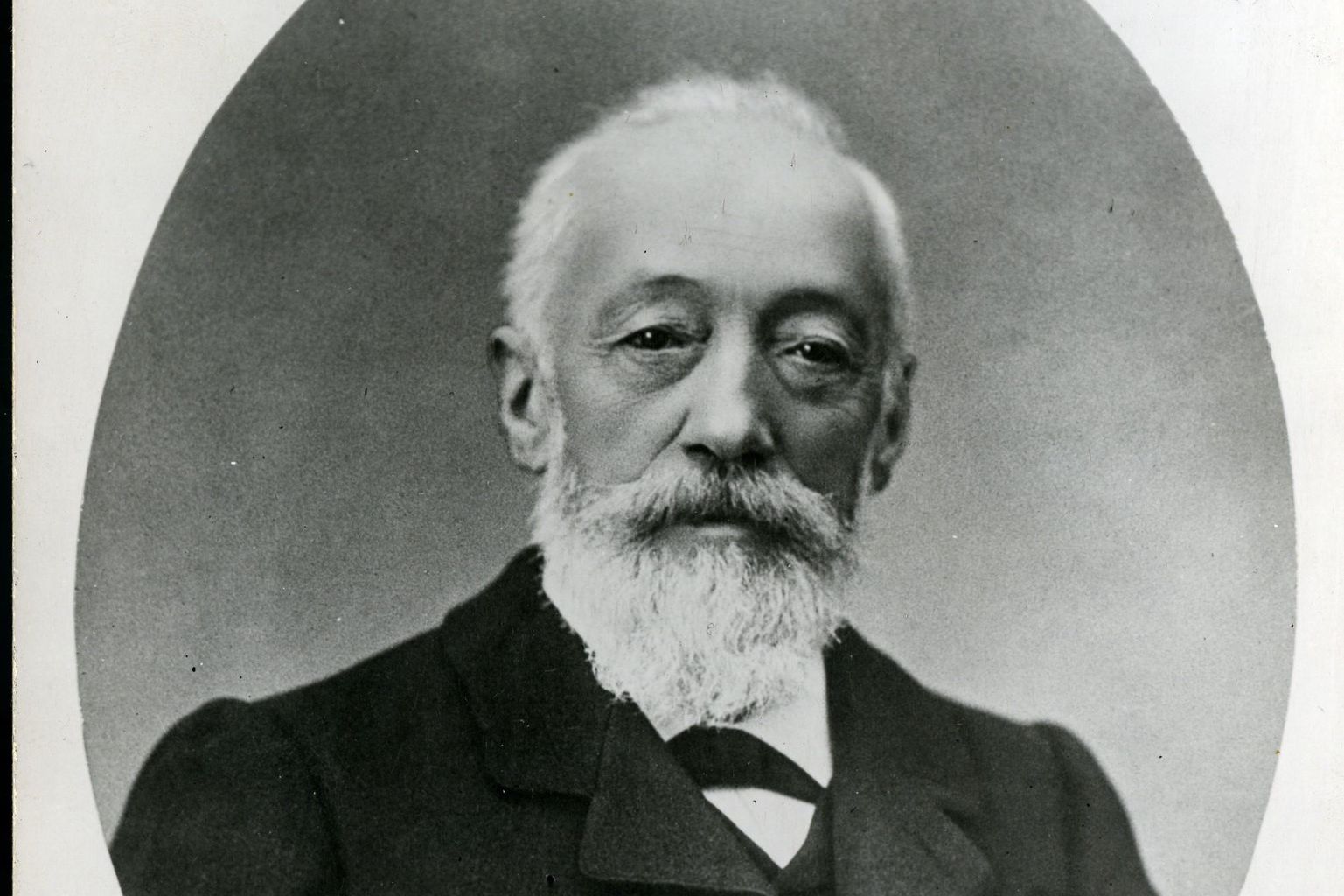 Portrait en noir et blanc d’un homme âgé avec une barbe, portant un costume sombre.