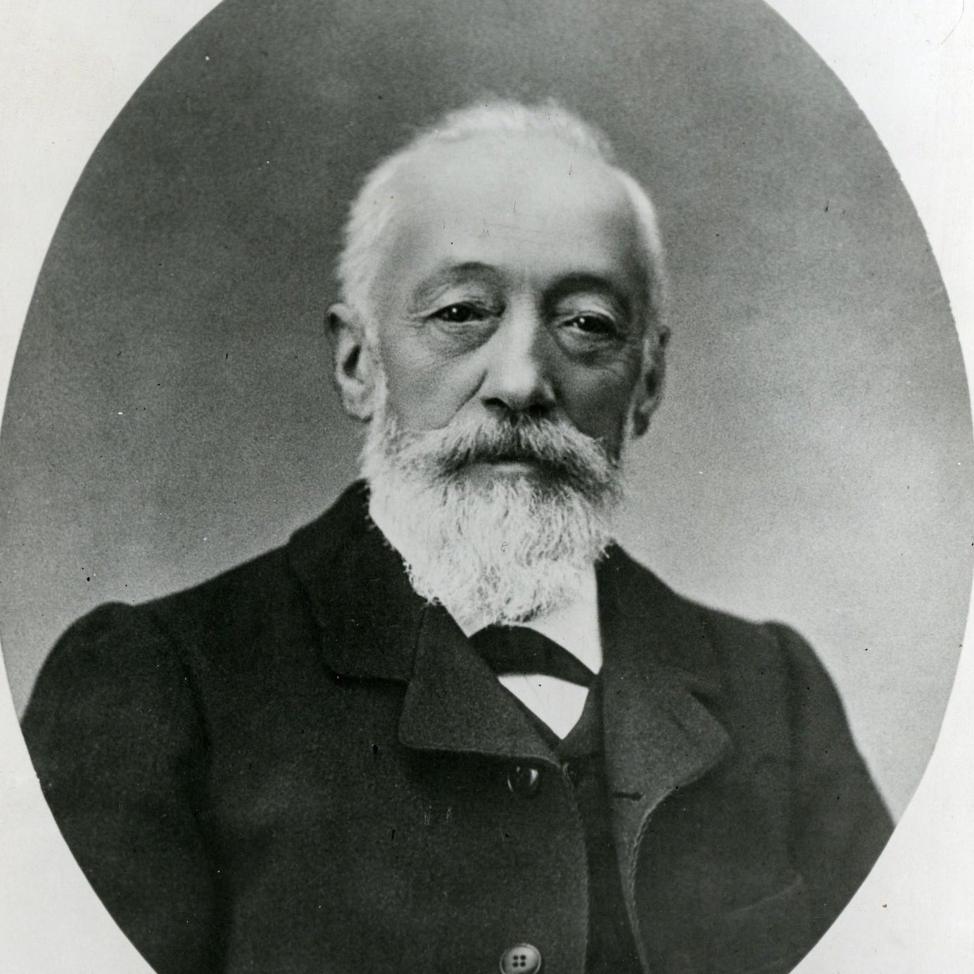 Portrait en noir et blanc d’un homme âgé avec une barbe, portant un costume sombre.