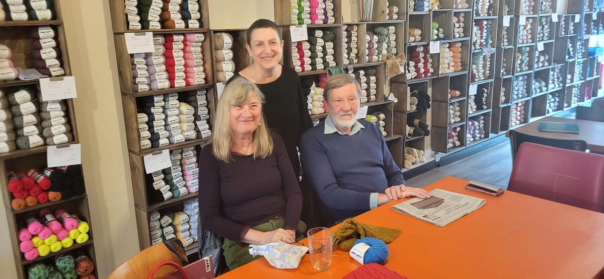 Trois personnes souriantes assises à une table dans un magasin de laine, entourées d'étagères remplies de fils colorés.