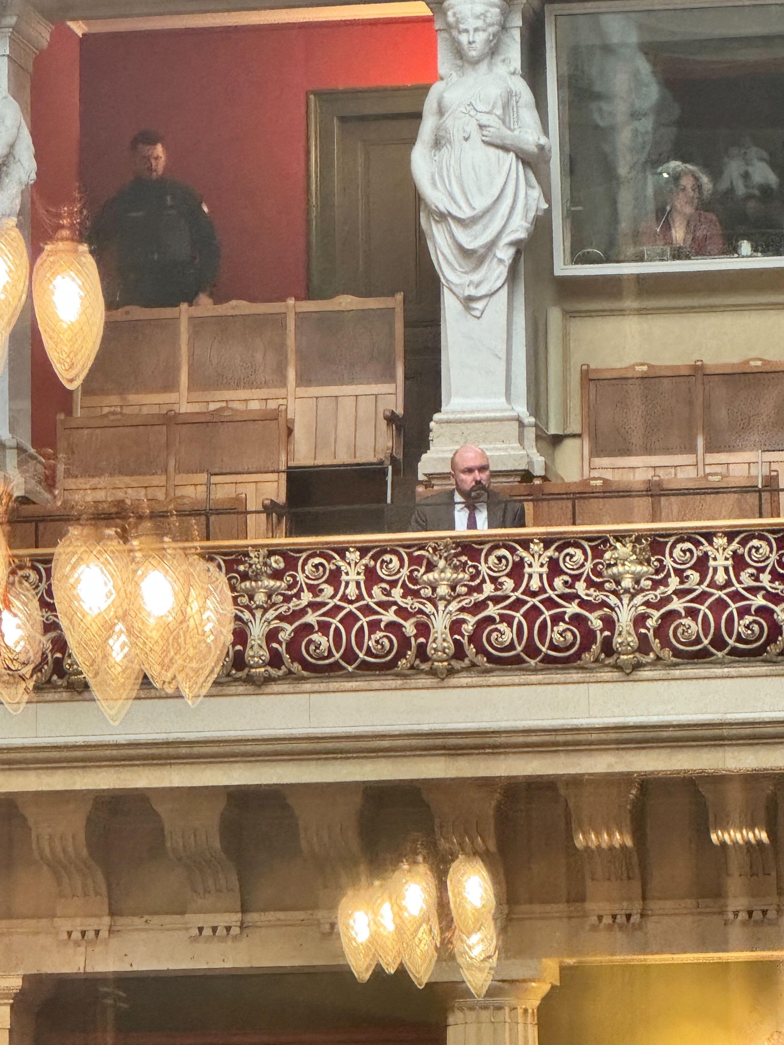 Eine kunstvoll verzierte Balkonbrüstung in einem historischen Gebäude, mit einem Sicherheitsbeamten im Hintergrund und einer sitzenden Person.