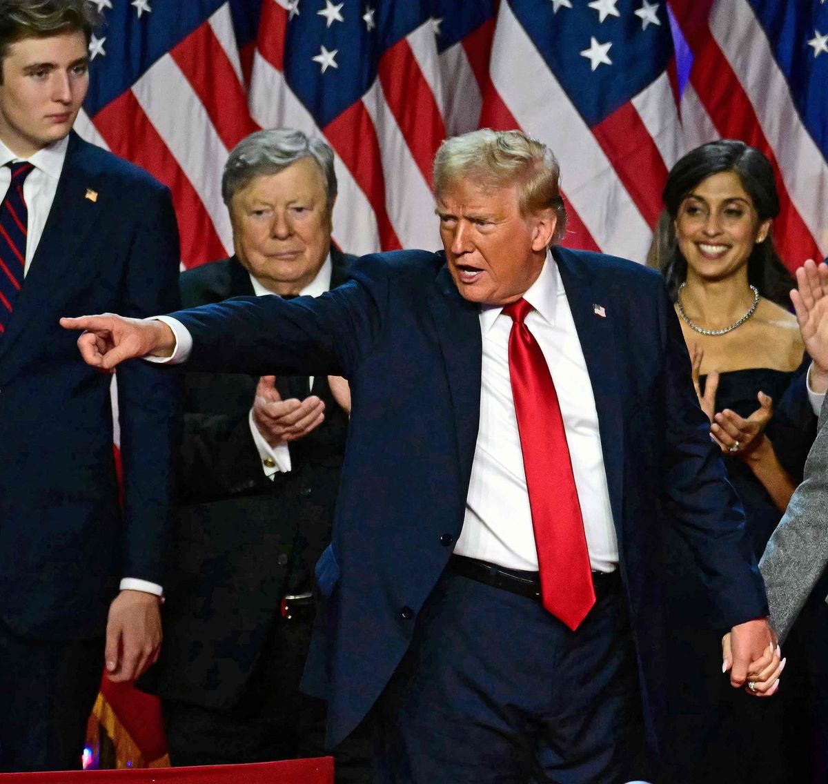 Donald Trump tenant la main de Melania Trump, faisant un geste vers ses partisans lors d'un événement électoral à West Palm Beach, devant des drapeaux américains, novembre 2024. AFP