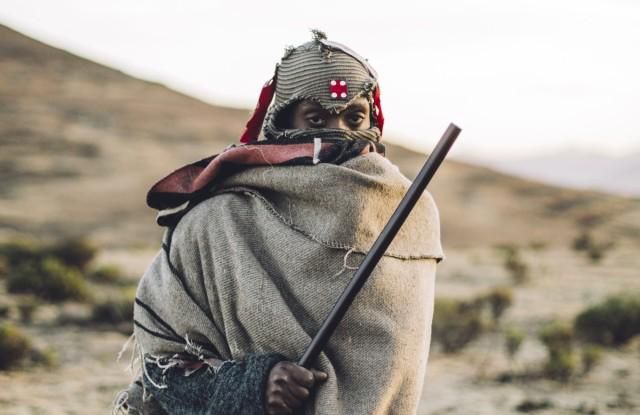 Person in traditioneller Kleidung mit Stab in einer kargen Landschaft bei Dämmerung.