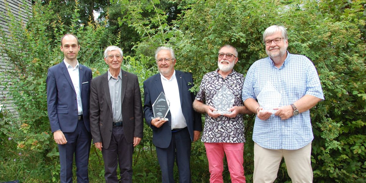 Cinq hommes en tenue formelle et décontractée posent côte à côte, tenant des trophées en verre transparent, devant un fond de buissons verts.