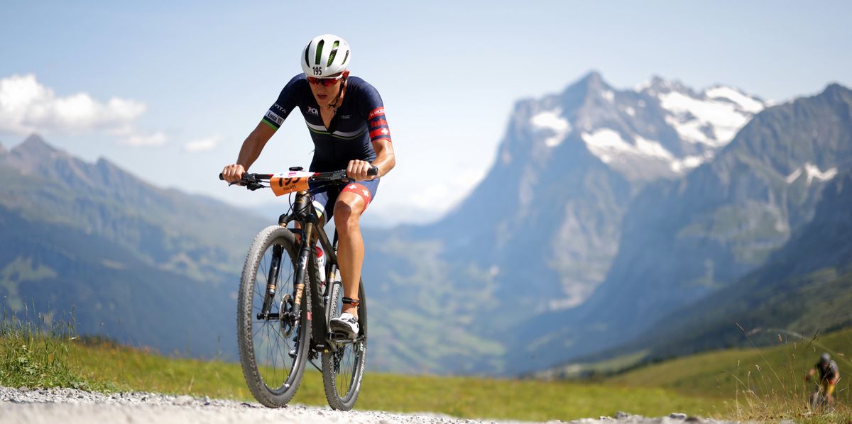 Ein Radfahrer fährt auf einem Schotterweg in einer bergigen Landschaft mit schneebedeckten Gipfeln im Hintergrund.