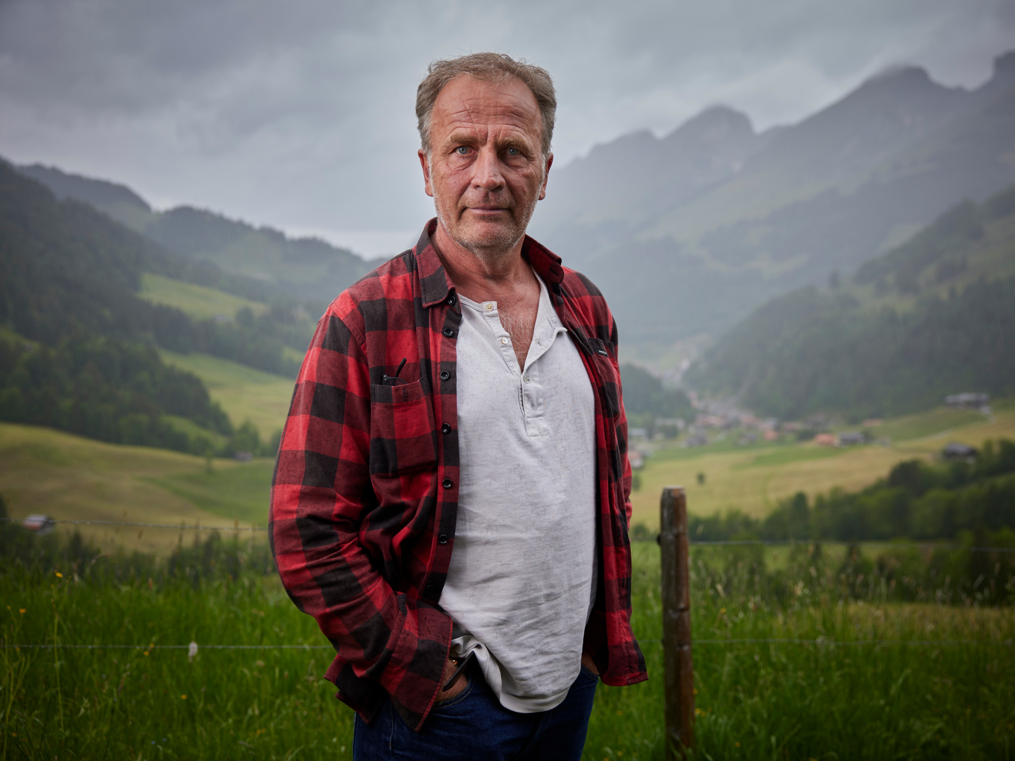 Schriftsteller Alex Capus steht am Jaunpass vor alpiner Bergkulisse, im rot-schwarzen Flanellhemd.
