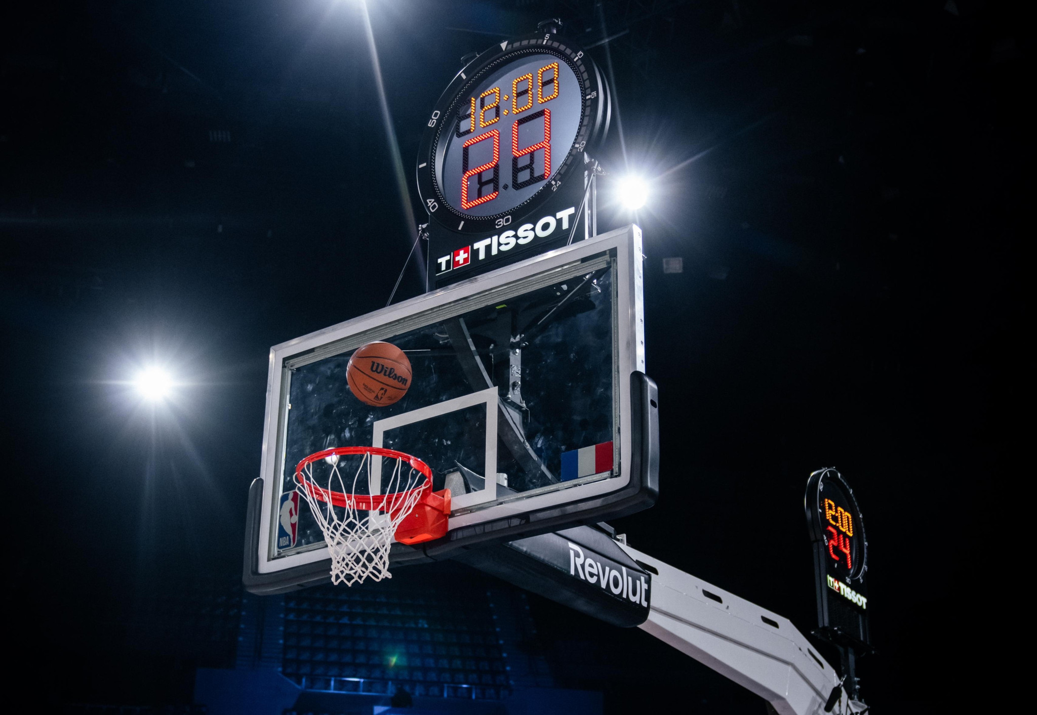Basketball in the air above the basket, with a digital tissot litter watch in the background in a sports arena.