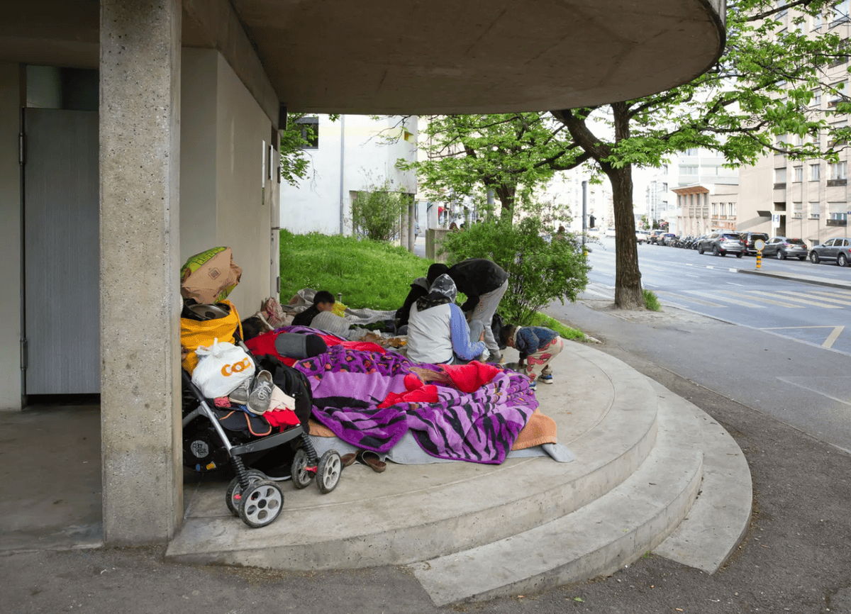 Groupe de personnes sans-abri dormant sous un abri en béton, avec des couvertures colorées et un chariot à provisions.