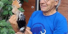 Un homme âgé portant des lunettes tient un oiseau huppé dans ses mains devant un bâtiment en bois avec des arbres en arrière-plan.
