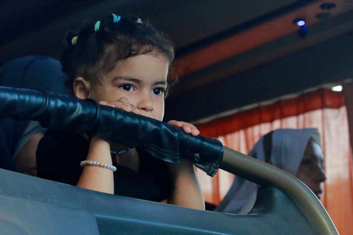Une jeune fille avec des cheveux bouclés regarde à travers une fenêtre tout en s'appuyant sur un dossier de siège, avec une femme en arrière-plan.