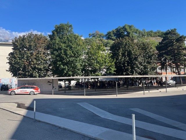 Parking de vélos couvert avec des arbres verts à côté d'un bâtiment. Une voiture de police est stationnée à gauche.