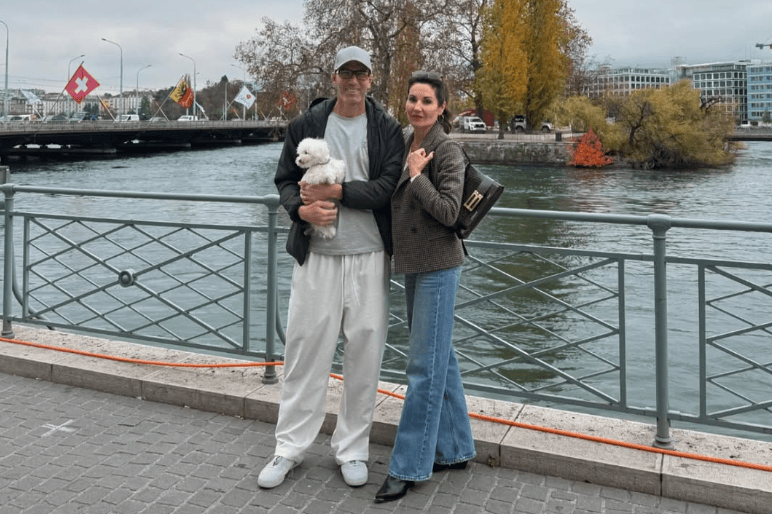 Un couple se tient sur une promenade au bord d’une rivière, près d’un pont. L’homme porte une casquette et des vêtements décontractés, tenant un petit chien blanc. La femme est habillée avec un manteau chic et un sac à main. Des drapeaux suisses sont visibles en arrière-plan, ainsi que des arbres d’automne.