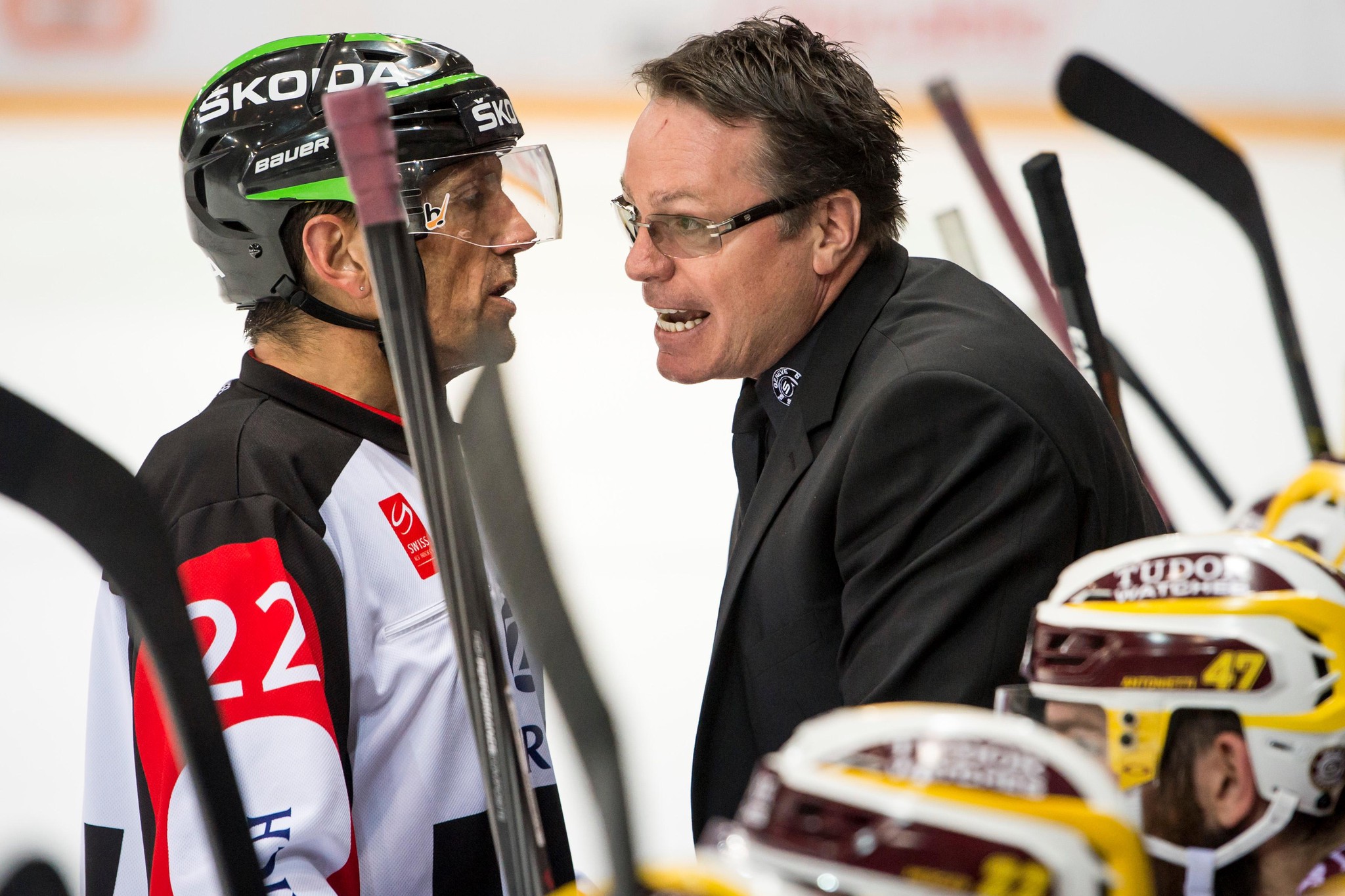 Eishockey-Trainer spricht energisch mit Schiedsrichter während eines Spiels. Spieler und Ausrüstung sind im Hintergrund zu sehen.