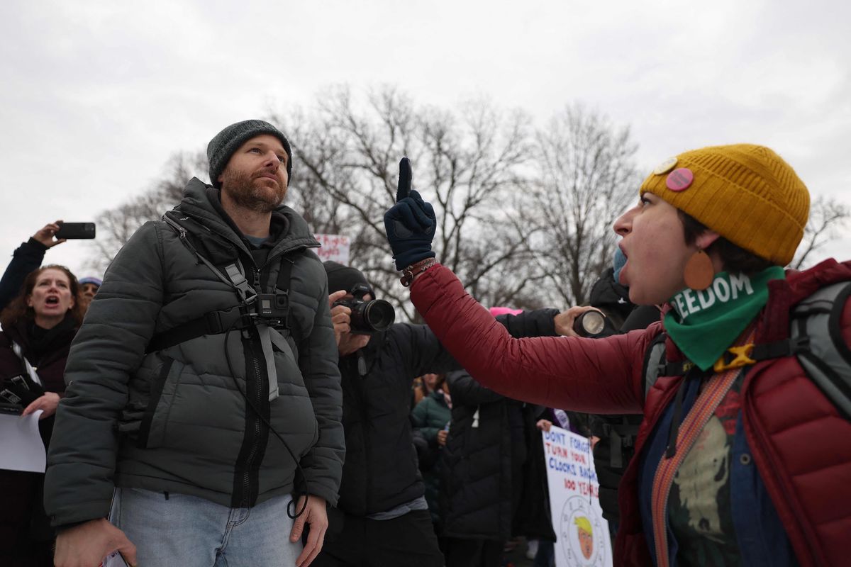 Un activiste gesticule en direction d’un autre participant lors de la marche ’People’s March on Washington’ à Washington, DC, le 18 janvier 2025, sous un ciel gris. Plusieurs participants sont visibles en arrière-plan, certains prenant des photos.