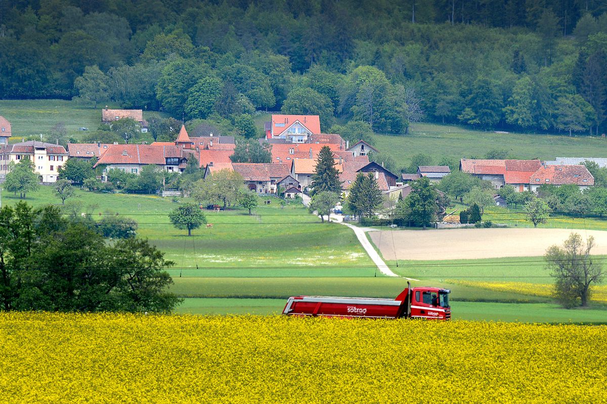 Vue du village de Berolle avec des maisons et des toits rouges entourés de verdure, et un tracteur rouge dans un champ de colza en premier plan.