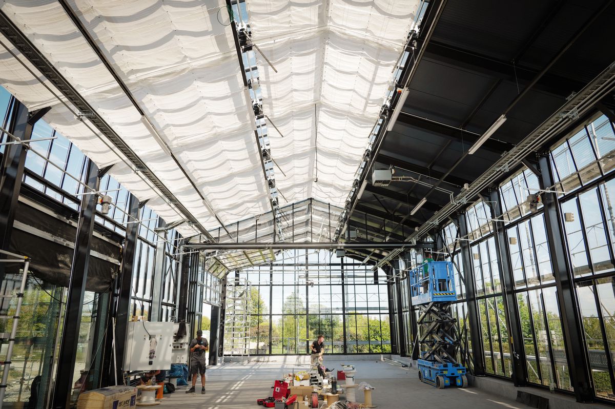 Intérieur d'un bâtiment en construction à Satigny, Genève, avec de grandes fenêtres et un plafond haut. Un chariot élévateur et des outils sont visibles au sol.