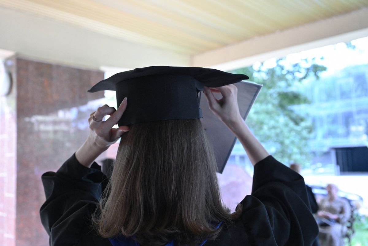 Une étudiante ajuste sa toque de graduation, tenant un diplôme, sous un auvent.