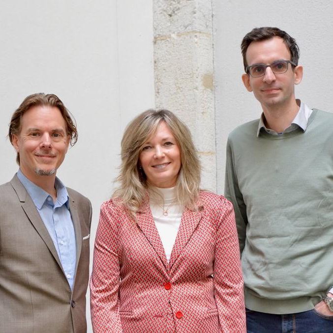 Trois personnes posent ensemble, deux hommes et une femme, tous souriants et bien habillés. L'homme à gauche porte un costume beige, la femme au centre un blazer rose, et l'homme à droite un pull vert.