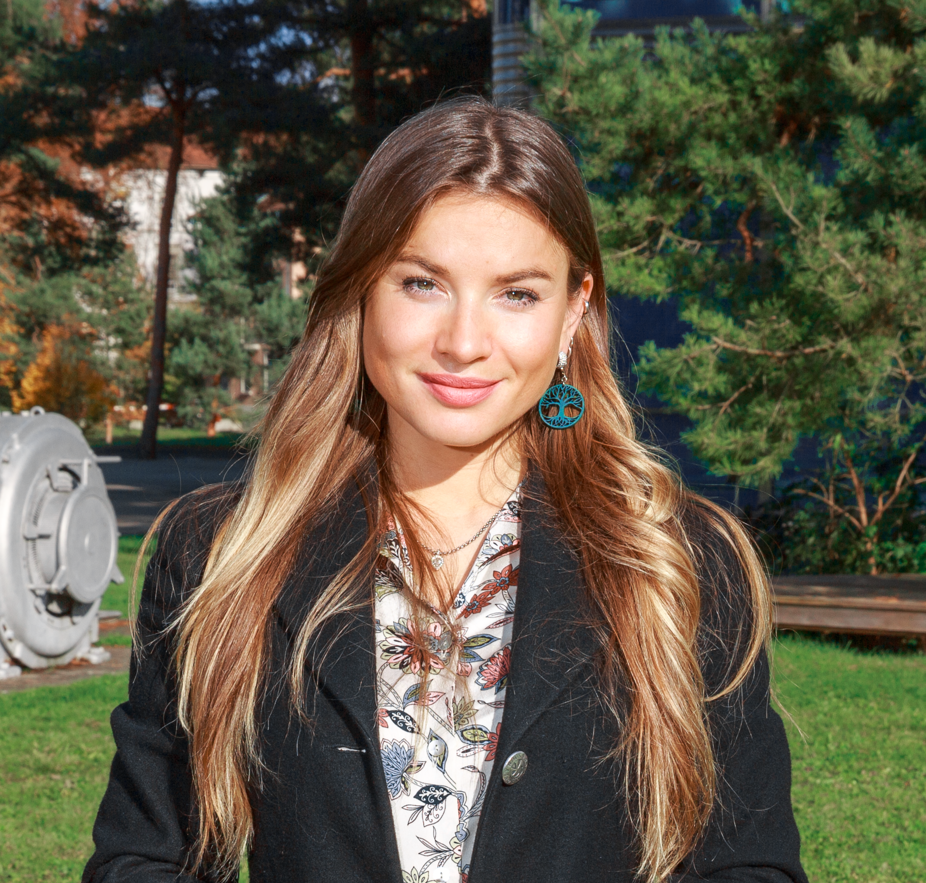 Junge Frau mit langen braunen Haaren und türkisfarbenen Ohrringen, schwarzem Blazer, vor grünen Bäumen im Sonnenlicht.