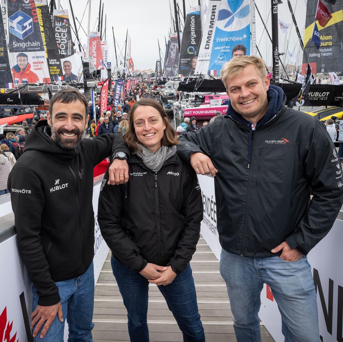 Trois navigateurs suisses, Alan Roura, Justine Mettraux et Oliver Heer, posent devant les bateaux du Vendée Globe aux Sables d’Olonne.