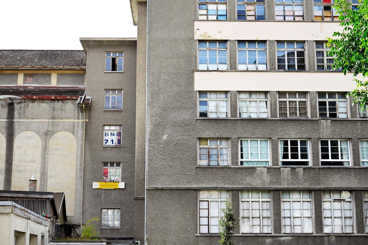 Bâtiment urbain gris à plusieurs étages avec de nombreuses fenêtres, certaines ouvertes. Arbre visible sur le côté droit.