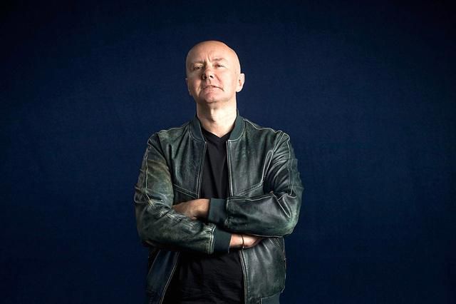 Personne portant une veste en cuir vert foncé, les bras croisés, posant devant un fond bleu foncé.