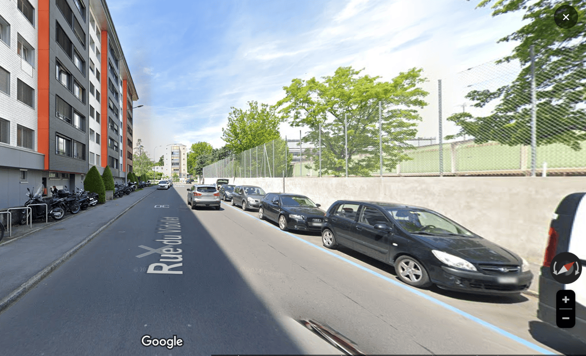 Rue urbaine à Genève avec des voitures garées le long du trottoir, bâtiments résidentiels à gauche et arbres à droite.