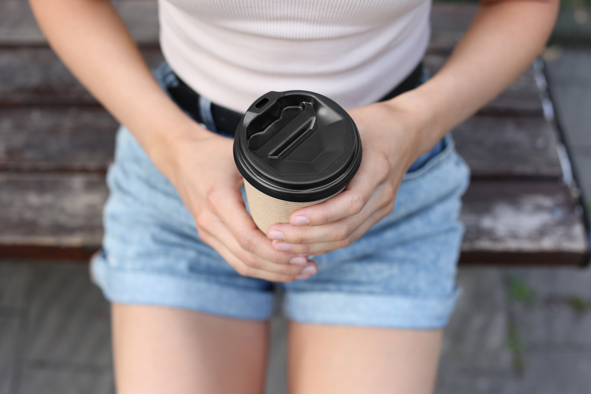 Frau in Jeansshorts hält einen Coffee-to-go-Becher aus Papier mit schwarzem Deckel im Freien.