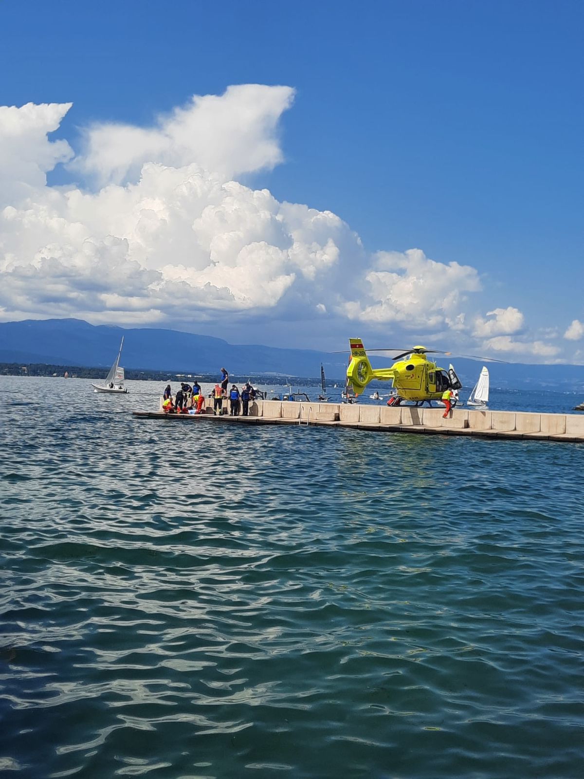 Un hélicoptère jaune est stationné sur un quai avec des personnes autour, à côté d'un lac avec des voiliers. Ciel partiellement nuageux en arrière-plan.