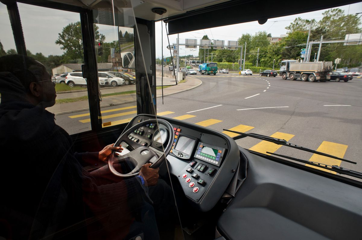 Vue depuis l'intérieur d'un trolleybus ligne 25 à Lausanne au carrefour de la Bourdonnette. Le conducteur tient le volant, et la route devant comporte plusieurs voies et véhicules, avec des panneaux de signalisation visibles en arrière-plan.
