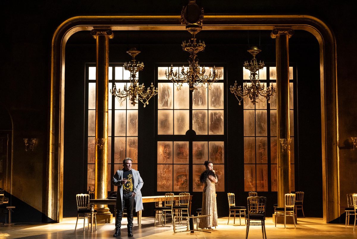 Scène d’opéra avec deux personnages sur scène, décor riche avec des chandeliers dorés et de grandes fenêtres.
